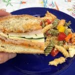 slice of picnic sandwich held in hand over blue plate with pasta salad