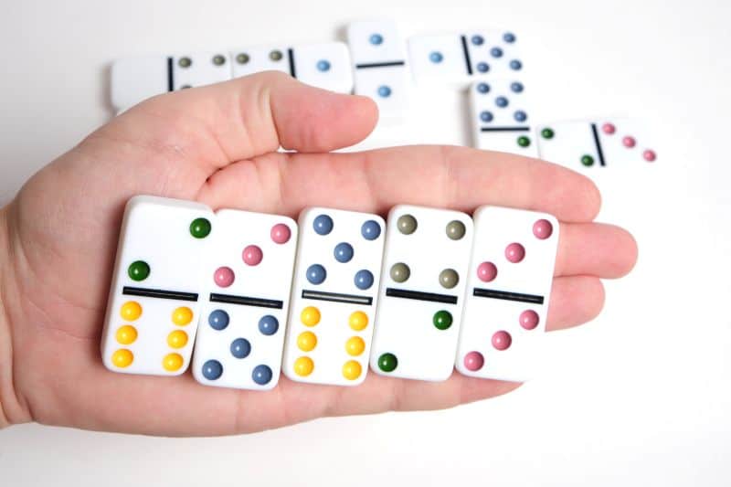 white hand holding white dominoes with colored pips over more white dominoes on white surface